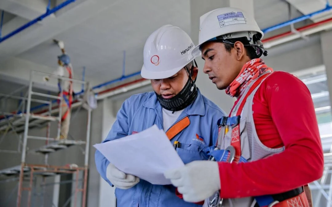 Two construction workers wearing safety helmets and gear are reviewing blueprints at a site, representing prefabrication in civil engineering.